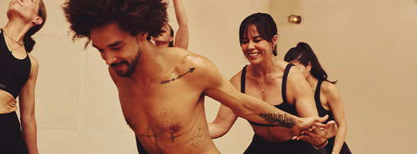 Group of smiling adults dancing together in a warm beige indoor setting, with arms linked and bodies captured in energetic motion