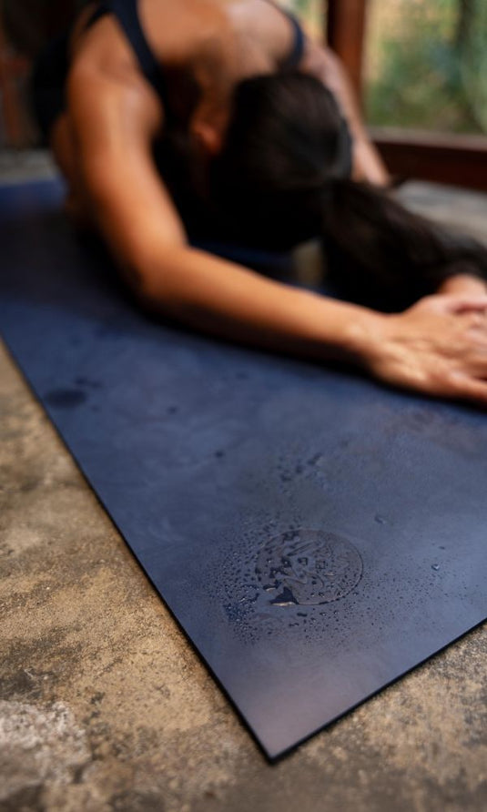 Person stretching forward on a dark blue Manduka yoga mat with an embossed logo, arms extended and hands resting near the front edge.