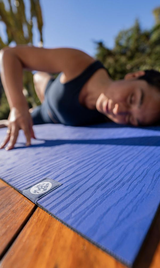 Woman practicing yoga outdoors on a blue Manduka yoga mat with a textured surface and visible logo in warm sunlight